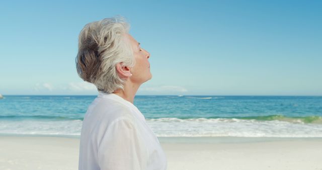 An elderly woman with short white hair stands on a quiet beach, basking in the peacefulness of the surrounding sea and sky. She gazes upwards, eyes closed, in a serene and meditative moment. This image is ideal for use in advertisements or articles about senior wellness, peaceful retirement, mindfulness, vacations, and enjoying nature.