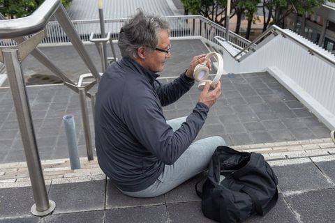 Senior asian man adjusting wireless headphones while resting on city steps with gym bag