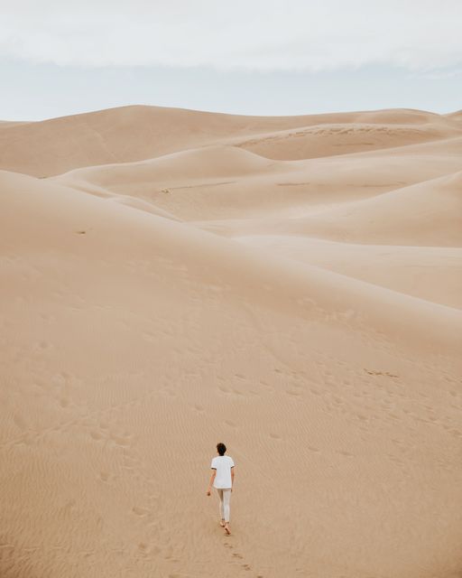 View of a person walking through vast sand dunes in a desert landscape. Ideal for use in projects related to nature, exploration, travel, solitude, or environmental concepts. Can be used in promotional materials for adventure tourism, travel guides, and educational content about deserts and natural landscapes.