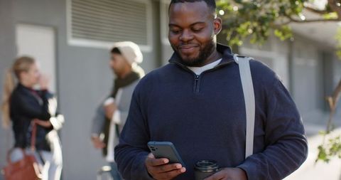 African american commuter checking phone while holding coffee on sunlit urban sidewalk