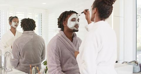African american couple wearing robes applying facial masks during at-home spa morning routine