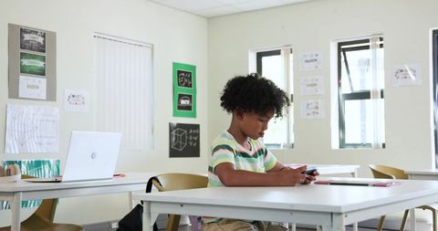 African American teen sitting in bright classroom using smartphone during independent study