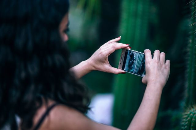 Woman taking close-up photo of green cactus outdoor using smartphone. Suitable for use in articles about hobbies, technology, nature photography, and modern lifestyle habits.