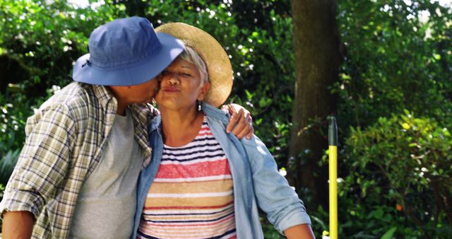 A middle-aged Asian man affectionately kisses a middle-aged Asian woman on the cheek in a lush garden setting, with copy space. Their casual attire and warm interaction suggest a moment of love and companionship in a serene outdoor environment.