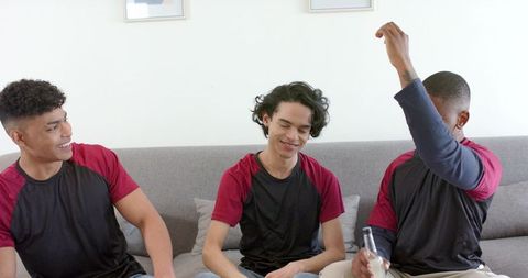 Young men celebrating together on couch wearing matching shirts, laughing and sharing drink