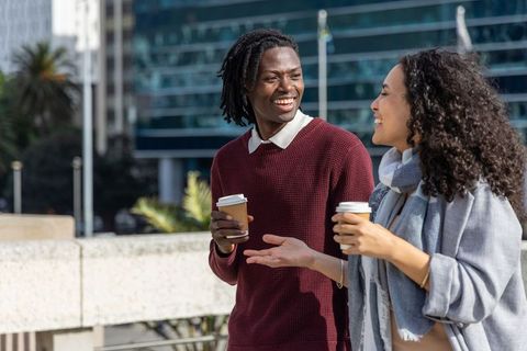 Young diverse friends walking urban street holding takeaway coffee cups, laughing together