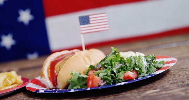 Hot dog on bun with ketchup, served with fresh salad on patriotic-themed plate, American flag in background. Ideal for content on American holidays such as Fourth of July, Memorial Day, and Independence Day. Perfect for food blogs, festive event promotions, and outdoor celebration advertisements.