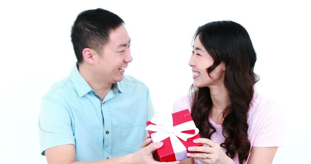 Couple exchanging a red gift box and smiling at each other. Ideal for use in content related to celebrations, relationship goals, special occasions like anniversaries or Christmas, and advertisements aimed at couples and gift products.