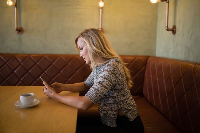 Woman sitting at a restaurant table using a mobile phone, with a cup of coffee in front of her. Ideal for illustrating concepts of modern lifestyle, technology use, social media engagement, and casual dining. Suitable for blogs, advertisements, and articles related to technology, communication, and leisure activities.
