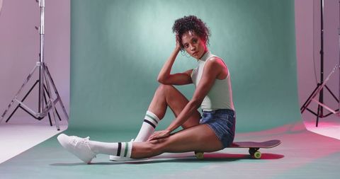 Woman posing on skateboard in pastel studio wearing tank top, denim shorts, knee socks
