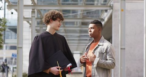 Multiracial graduates conversing on modern campus walkway holding cap and diploma