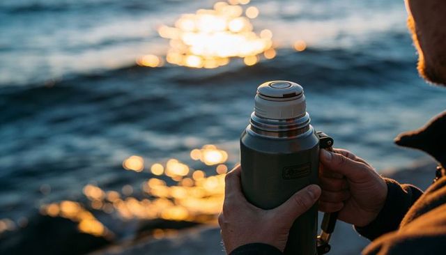 Holding thermos at sunset sea, twisting lid on rocky coastline during golden hour
