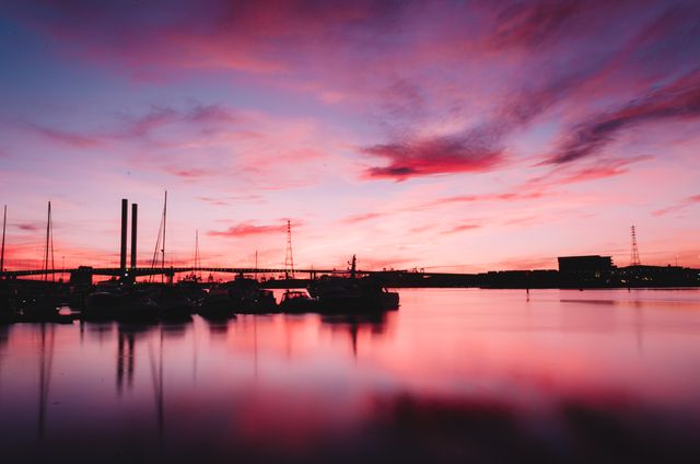 Silhouettes of boats anchored at a dock with a bridge in the background set against a vibrant sunset with pink and purple tones. Calm waters gently reflect the colors of the sky. Suitable for backgrounds, travel brochures, social media posts, or advertisements emphasizing serenity and beauty.