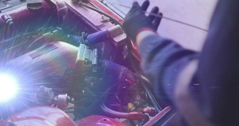 Mechanic repairing engine bay with black-gloved hand and exposed wiring under lens flare