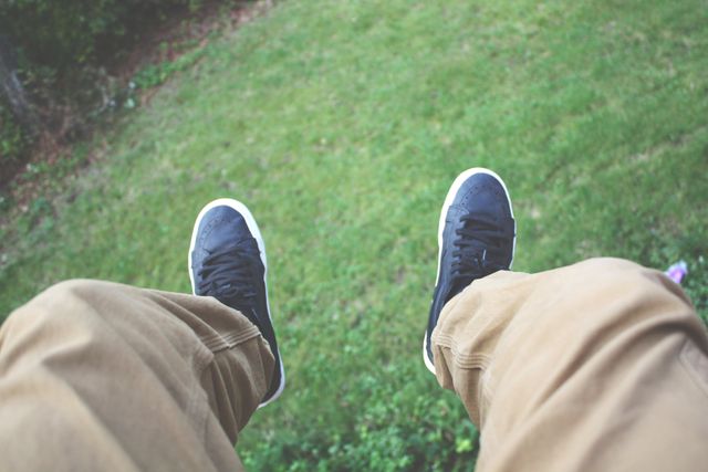 Person wearing khaki pants and black sneakers relaxing outdoors, with legs dangling over a green grassy field. Great for illustrating leisure, casual fashion, and an outdoor lifestyle. Can be used for blog posts, lifestyle advertisements, or social media content related to relaxation, adventure, or enjoying nature.