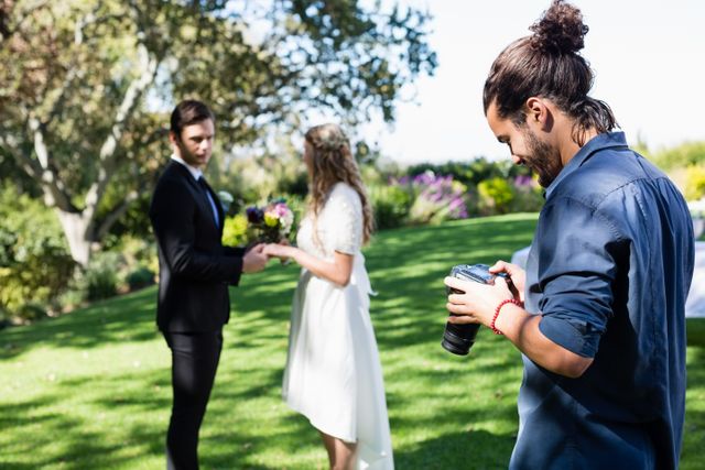 Photographer reviewing photos on digital camera while bride and groom exchange vows in park. Ideal for use in wedding planning, photography services, outdoor ceremonies, and event documentation.