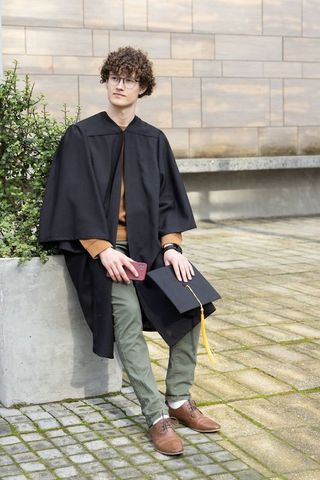 Contemplative young graduate sitting in gown holding mortarboard with yellow tassel