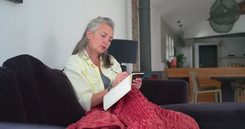 Senior woman writing in notebook on sofa, using phone, red blanket, cozy open-plan home
