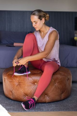 Woman tying running shoes on ottoman in modern living room preparing for morning workout