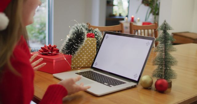 A person engages in a video call on a laptop surrounded by festive Christmas decorations, including wrapped presents, a miniature Christmas tree, and holiday ornaments. This can be used for advertisements and articles about staying connected with family during the holidays, virtual celebrations, or holiday-themed technology usage.