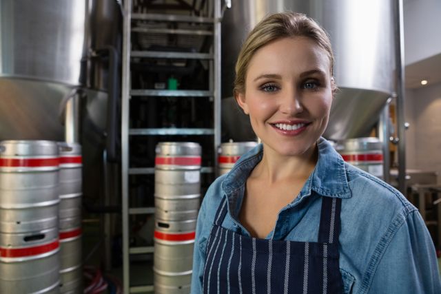This image shows a smiling female worker standing in front of storage tanks at a brewery. She is wearing a denim shirt and a striped apron, indicating her role in the beer production process. This image can be used for promoting the brewing industry, showcasing women in manufacturing roles, or highlighting professional environments in industrial settings.