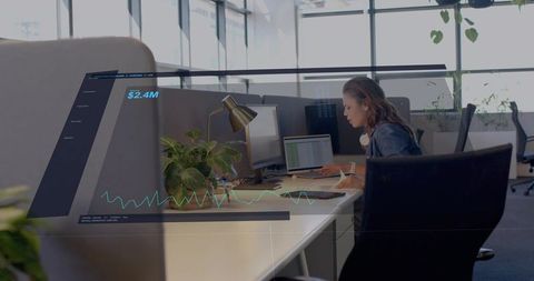 Businesswoman analyzing financial dashboard on laptop and monitor in modern open office