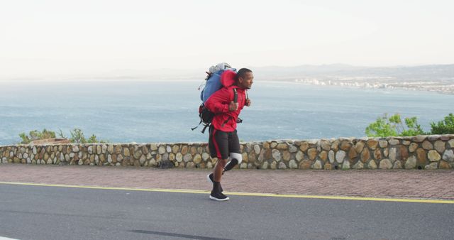 Determined biracial man with prosthetic leg trekking with backpack on coastal road. Long distance walking, fitness, challenge, disability, nature and healthy outdoor lifestyle.