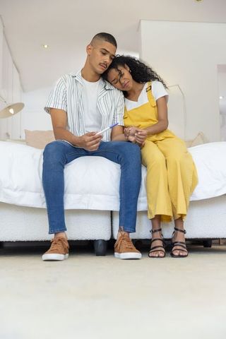 Young couple checking pregnancy test, holding hands on bed, woman leaning on partner