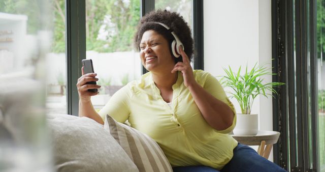 African American woman sitting on couch, joyfully listening to music on her smartphone using headphones. Natural light fills the room, and she appears relaxed and happy. This may be used for concepts related to technology, relaxation, leisure activities at home, enjoyment of music, or interior lifestyle.