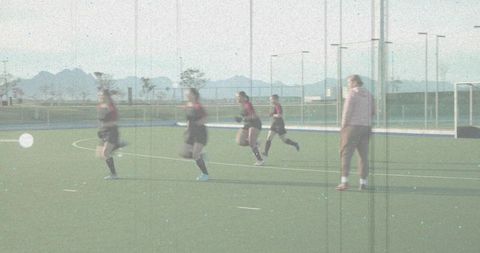 Female field hockey players sprinting across astroturf while coach observing training session