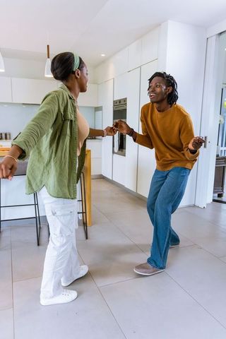 African american couple dancing in sunlit modern open-plan kitchen holding hands, playful energy
