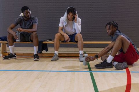 Multiracial male athletes resting on bench in gym holding water bottles and smartphone