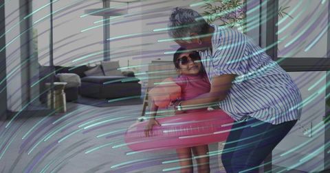 Woman hugging and adjusting child in pink swimsuit with inflatable swim ring in living room