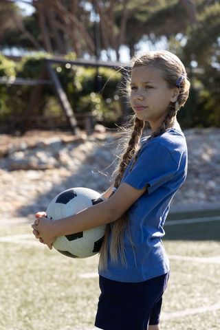 Young girl holding soccer ball on sunny turf wearing blue kit and braids