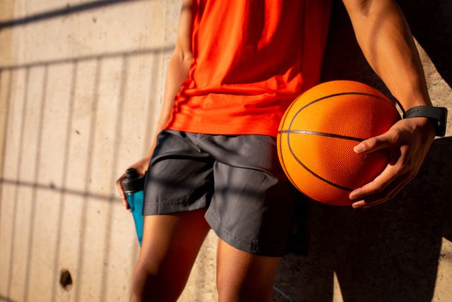 Midsection of fit biracial man holding basketball. sports, healthy active lifestyle and outdoor fitness.
