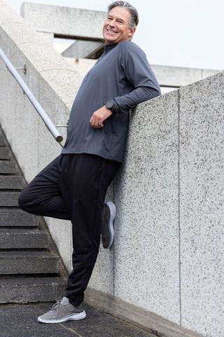 Smiling mature man leaning on concrete wall after urban workout wearing joggers and watch