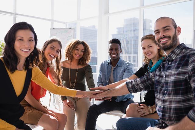 This image shows a diverse group of professionals forming a hand stack in a bright, modern office. They are smiling and appear to be engaged in a team-building activity, symbolizing unity and collaboration. This image is ideal for use in corporate presentations, team-building workshops, diversity and inclusion campaigns, and business websites to emphasize teamwork, cooperation, and a positive work environment.