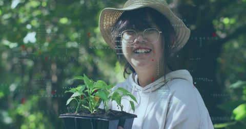 Smiling girl holding seedling tray wearing straw hat and glasses in sunlit garden hud