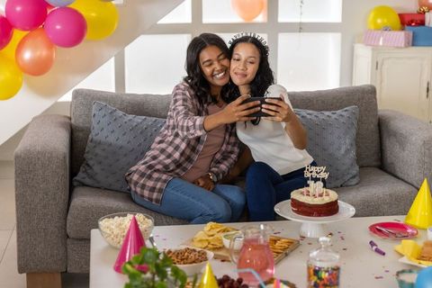 Diverse mother and teen daughter taking selfie on couch at birthday party