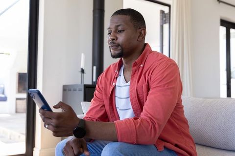 African american man sitting on sofa in modern living room holding smartphone and browsing