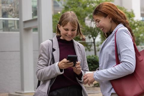 Two women laughing while checking smartphone outdoors on urban campus