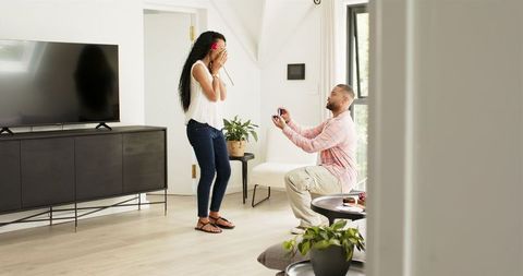 Man Proposing with Engagement Ring to Surprised Woman Holding Rose at Home