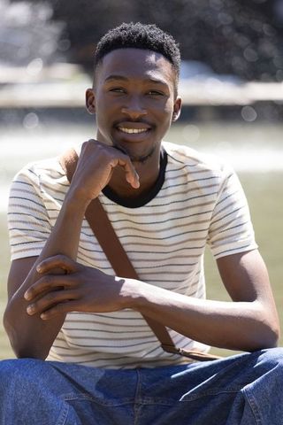 Young man smiling outdoors sitting by fountain in casual striped shirt and crossbody bag