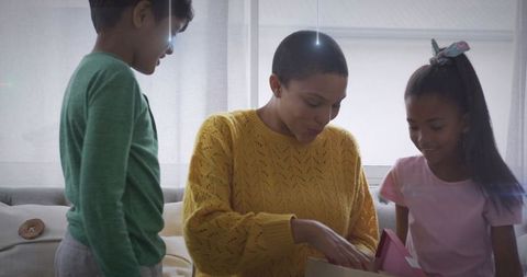Mother opening gift with children on sofa in sunlit living room, warm cozy family moment