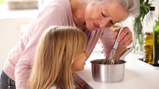 Grandmother showing granddaughter how to make chocolate sauce, sharing family recipe. Perfect for content on family bonding, cooking tutorials, home life, and generational learning. Can be used in articles about fostering family traditions, recipes, or educational materials on cooking.