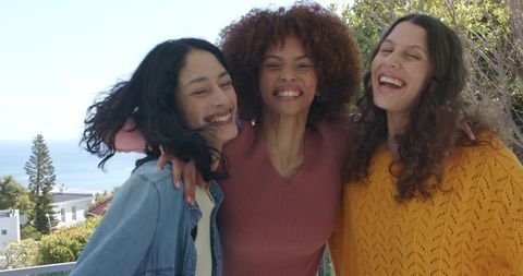 Multicultural women laughing and hugging on sunny seaside balcony overlooking ocean view