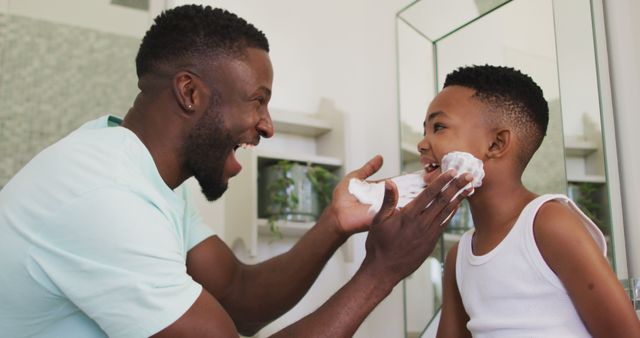 African american father putting shaving cream on his son mouth and laughing together. staying at home in self isolation during quarantine lockdown.