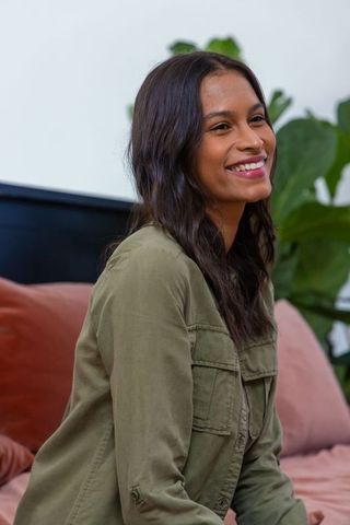 African american woman smiling on cozy bedroom bed wearing olive utility shirt with plants