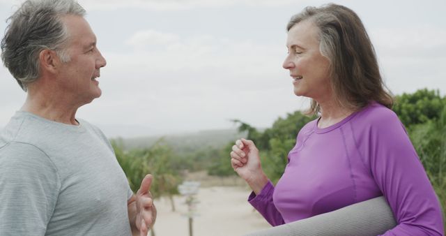 Senior couple engaging in conversation while standing outdoors and holding a yoga mat. They appear to be relaxed, enjoying fresh air and fitness activities. This image is ideal for promoting healthy lifestyles for seniors, advertising fitness programs for older adults, or showcasing positive, active aging.