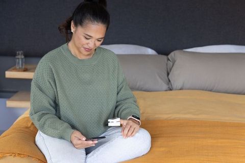 Middle-aged asian woman sitting on bed checking pulse oximeter and using smartphone, calm
